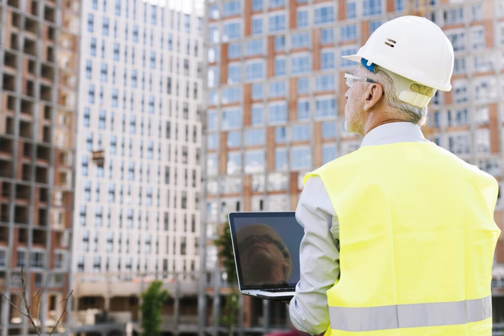 Construction site supervisor wearing a hard hat and high-visibility vest, using a laptop to inspect a high-rise building project.