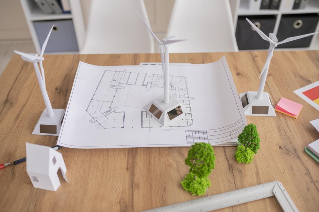 Architectural blueprint with wind turbine models and miniature trees on a wooden desk.