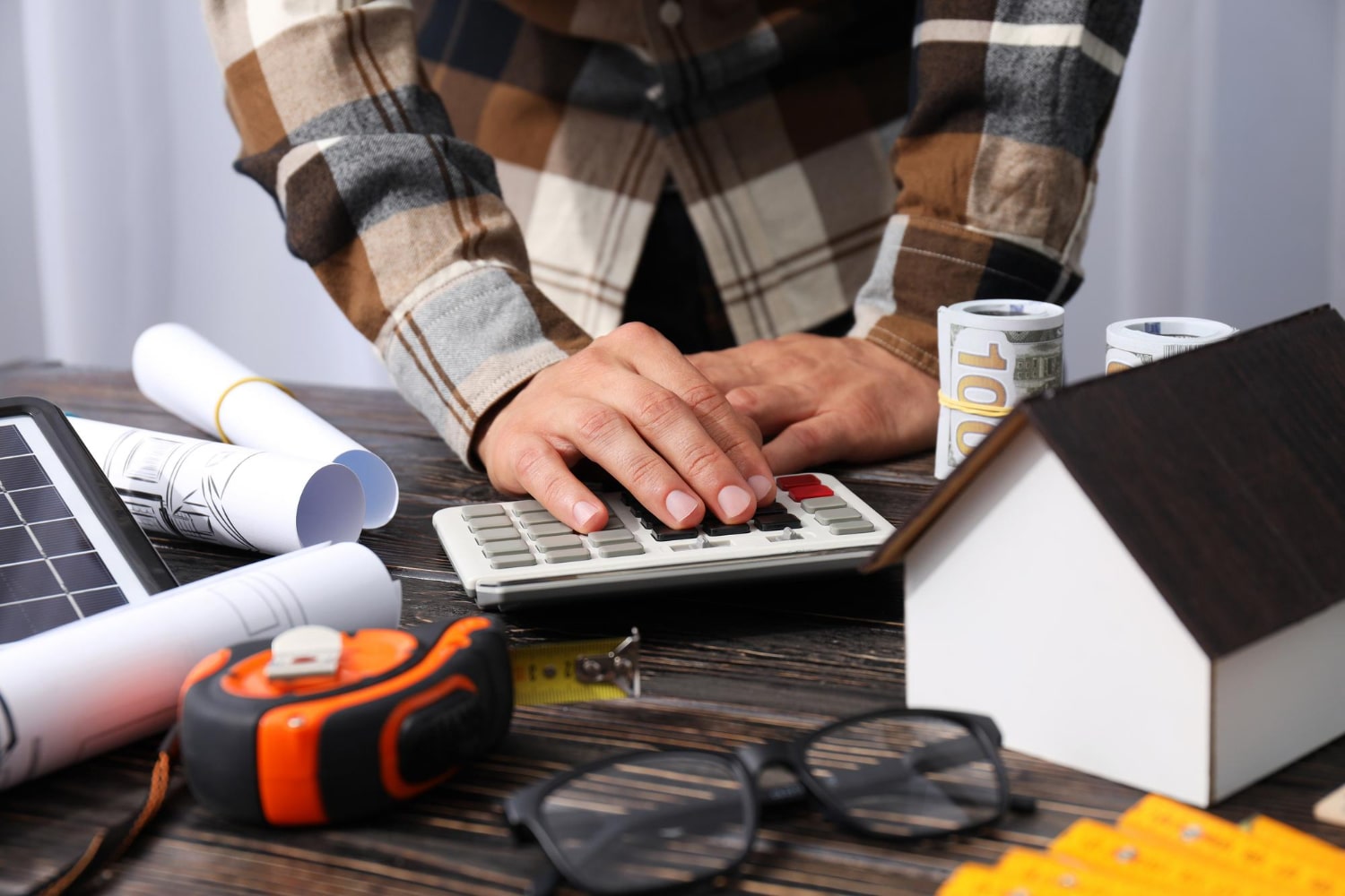 Hands using a calculator beside house model, blueprints, tools, and rolled banknotes, illustrating construction firm UK cost planning and budgeting.